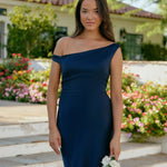A woman stands in front of a white back drop facing the back wearing a dark blue bridesmaid dress. This dress has an asymmetrical neckline and gathers slightly at the waist. Perfect as a bridesmaid dress or a special event dress. This is the Savannah Dress in Navy.