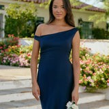 A woman stands in front of a white back drop facing the back wearing a dark blue bridesmaid dress. This dress has an asymmetrical neckline and gathers slightly at the waist. Perfect as a bridesmaid dress or a special event dress. This is the Savannah Dress in Navy.