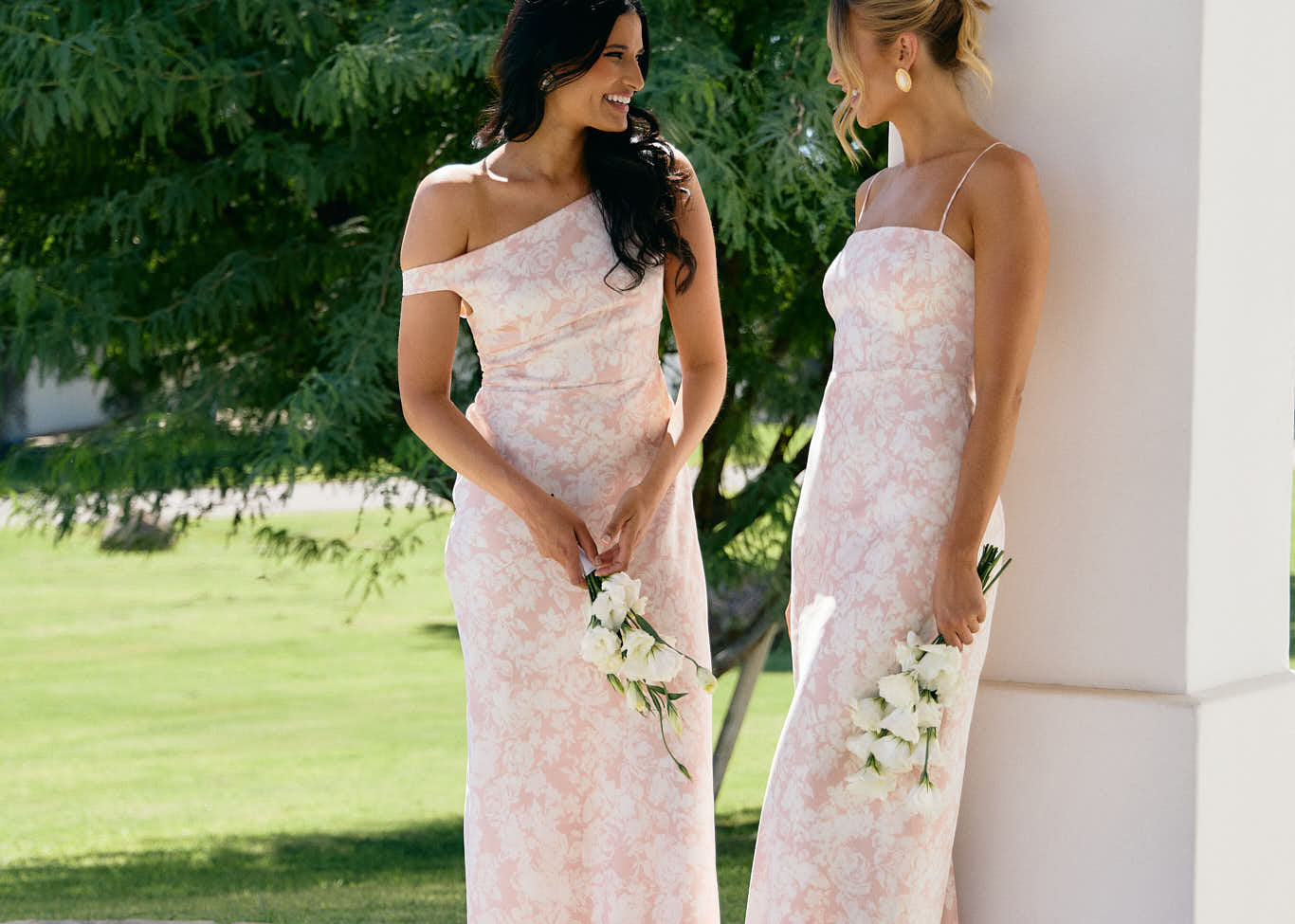 A woman stands in front of a white back drop wearing a light pink and white floral print bridesmaid dress. This dress has an asymmetrical neckline and gathers slightly at the waist. Perfect as a bridesmaid dress or a special event dress. This is the Savannah Dress in Alba.