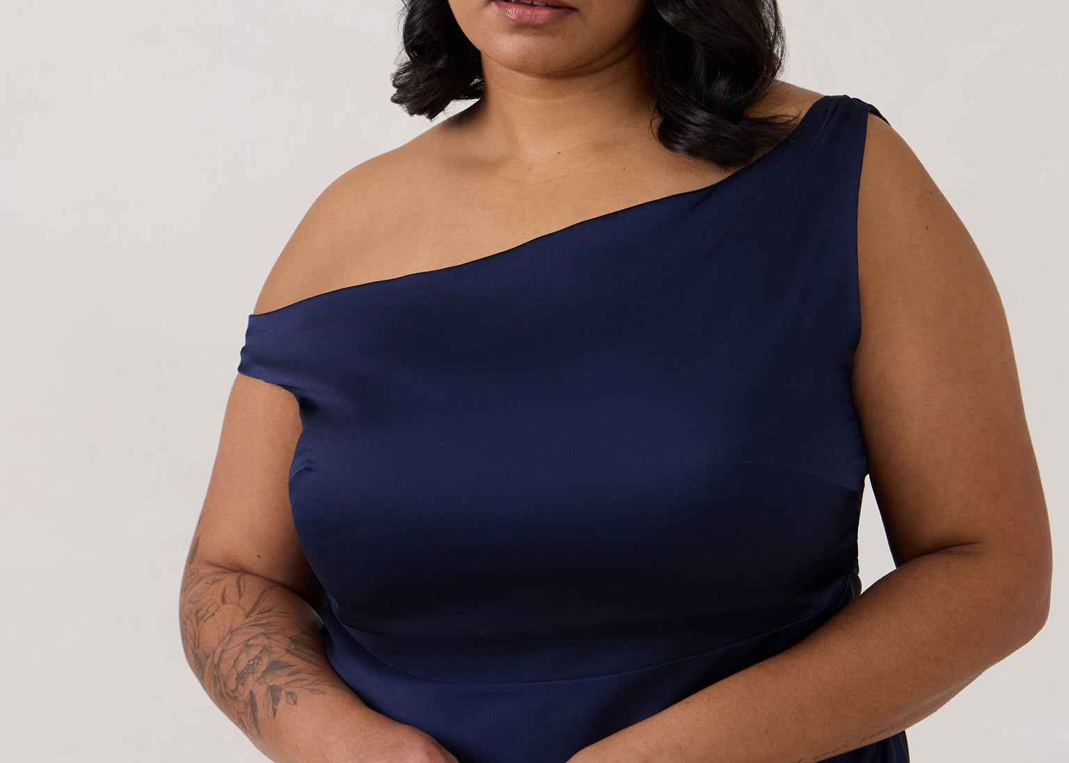 A woman stands in front of a white back drop wearing a dark blue plus size bridesmaid dress. This dress has an asymmetrical neckline and gathers slightly at the waist. Perfect as a bridesmaid dress or a special event dress. This is the Savannah Dress in Navy.