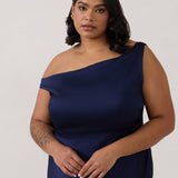 A woman stands in front of a white back drop wearing a dark blue plus size bridesmaid dress. This dress has an asymmetrical neckline and gathers slightly at the waist. Perfect as a bridesmaid dress or a special event dress. This is the Savannah Dress in Navy.
