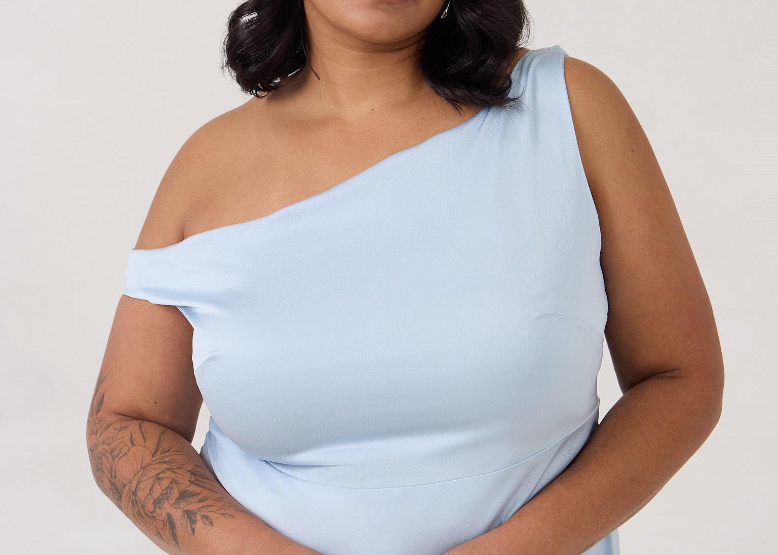 A woman stands in front of a white back drop wearing a light blue plus size bridesmaid dress. This dress has an asymmetrical neckline and gathers slightly at the waist. Perfect as a bridesmaid dress or a special event dress. This is the Savannah Dress in Powder Blue.