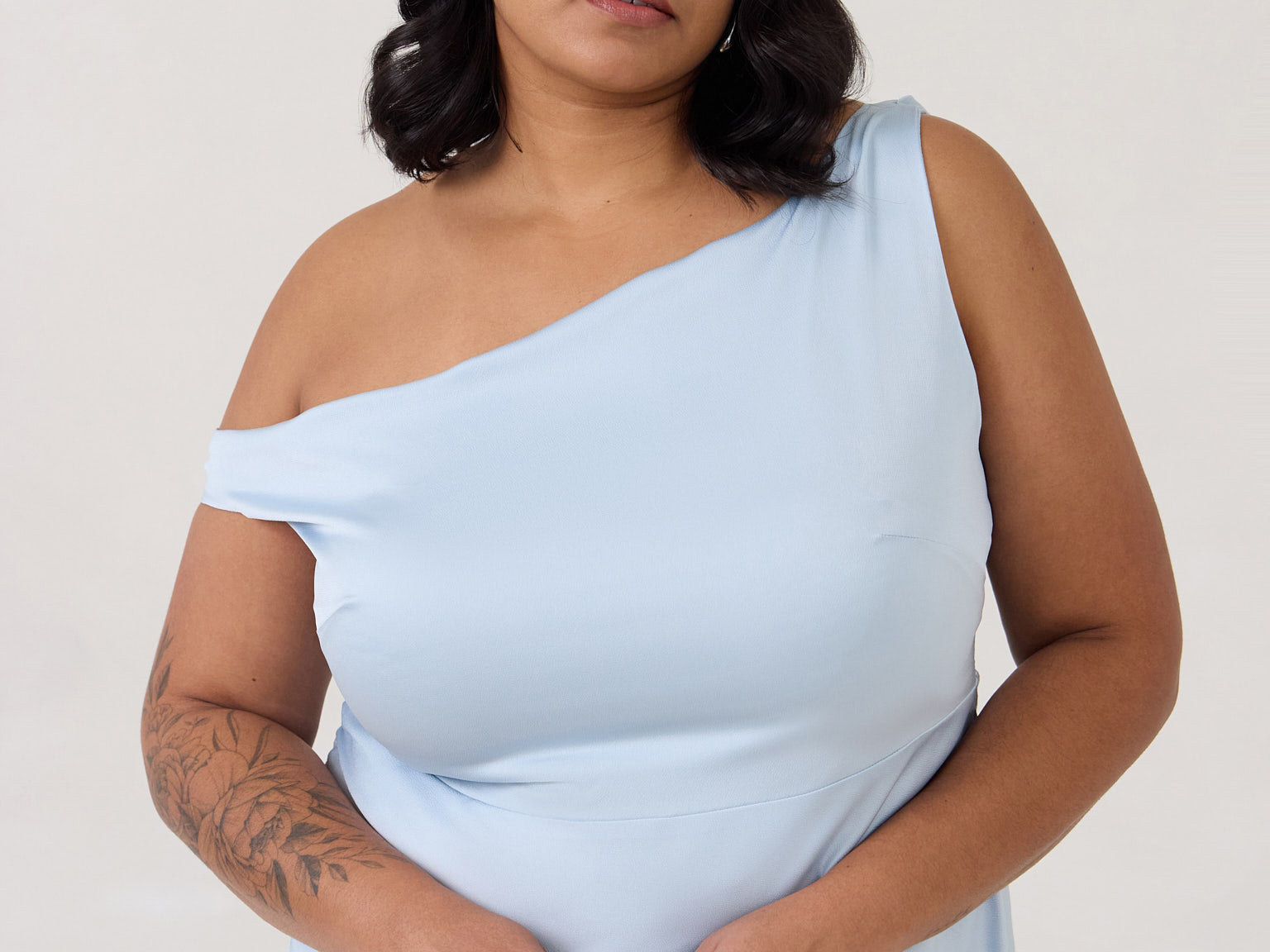 A woman stands in front of a white back drop wearing a light blue plus size bridesmaid dress. This dress has an asymmetrical neckline and gathers slightly at the waist. Perfect as a bridesmaid dress or a special event dress. This is the Savannah Dress in Powder Blue.
