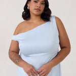 A woman stands in front of a white back drop wearing a light blue plus size bridesmaid dress. This dress has an asymmetrical neckline and gathers slightly at the waist. Perfect as a bridesmaid dress or a special event dress. This is the Savannah Dress in Powder Blue.